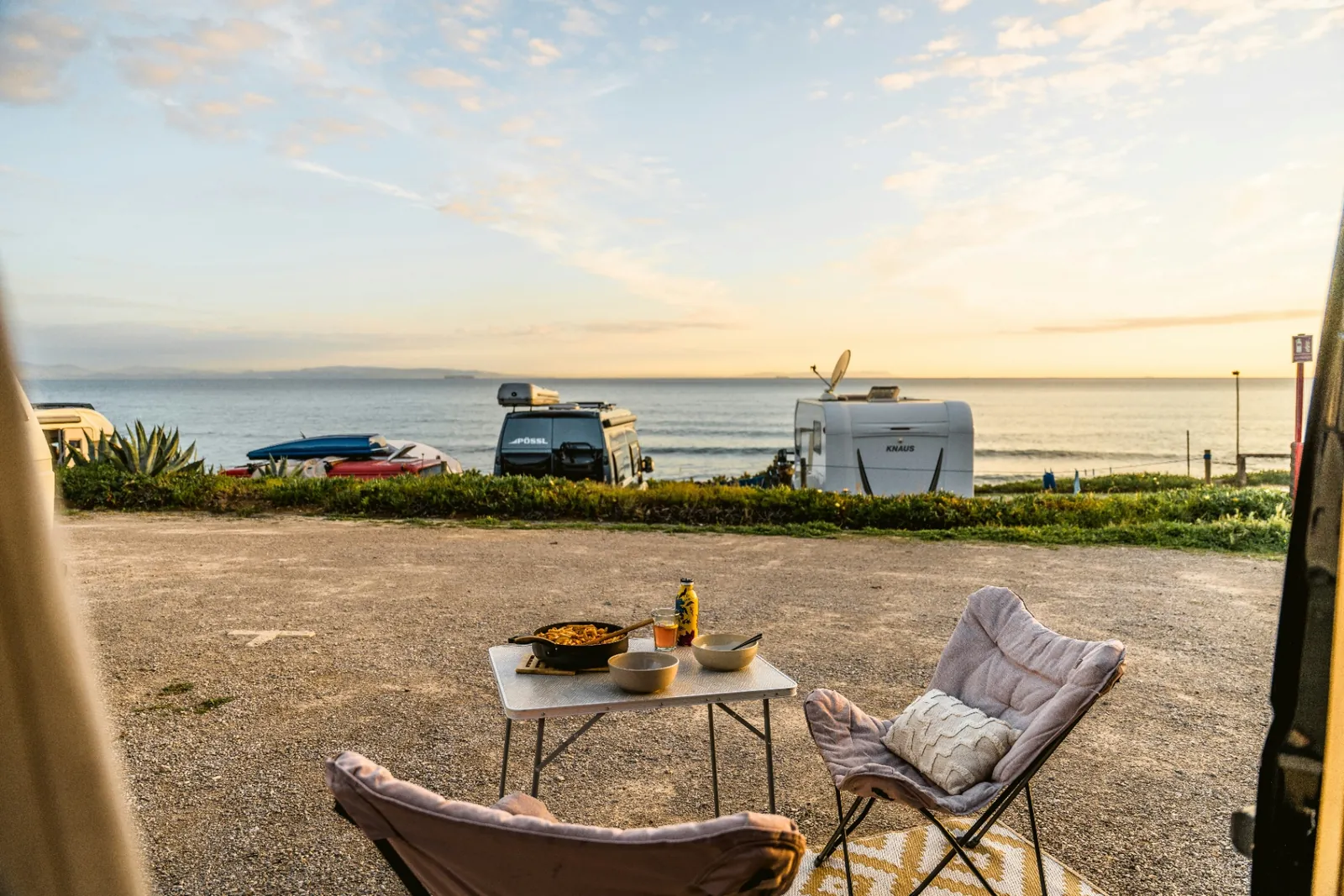 Camp chairs and a skillet of pasta on a folding table overlooking the ocean at sunset, with camper vans parked along the bluff