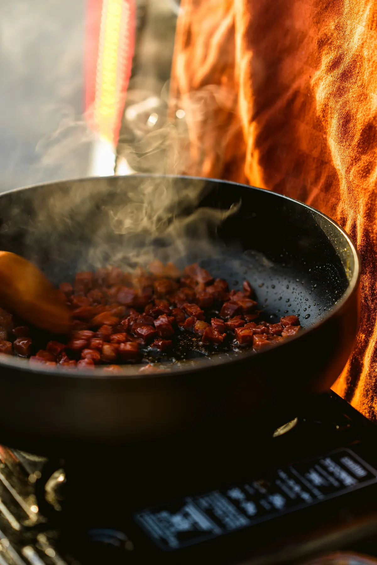 Diced chorizo sizzling in a black skillet over a camp stove at golden hour